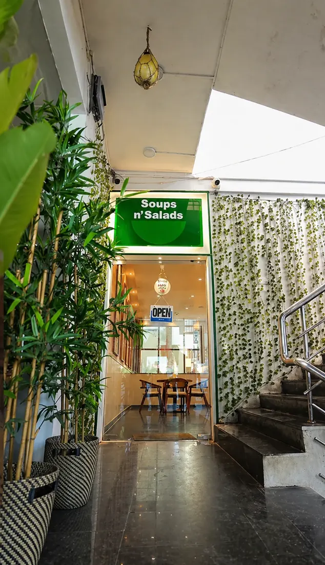The entrance to a small restaurant — a glass doorway under a lit sign, framed by plants, with a staircase leading up to the dining room